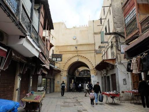 A street in the Mellah of Fes showing the characteristic archway that marks the transition between the wider medina and the Jewish quarter — commercial activity continues on both sides of the threshold